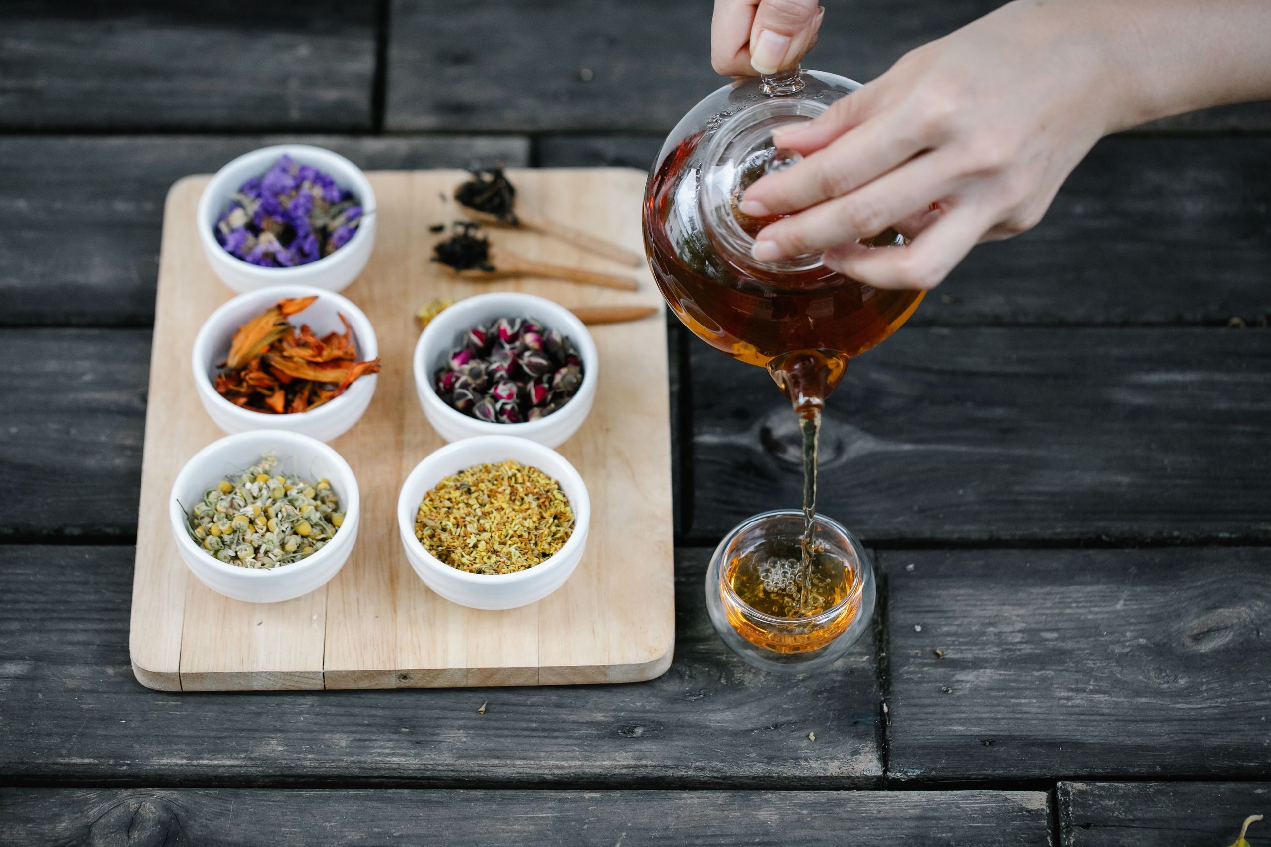A hand pouring herbal tea into a cup, surrounded by various dried herbs on a cutting board.