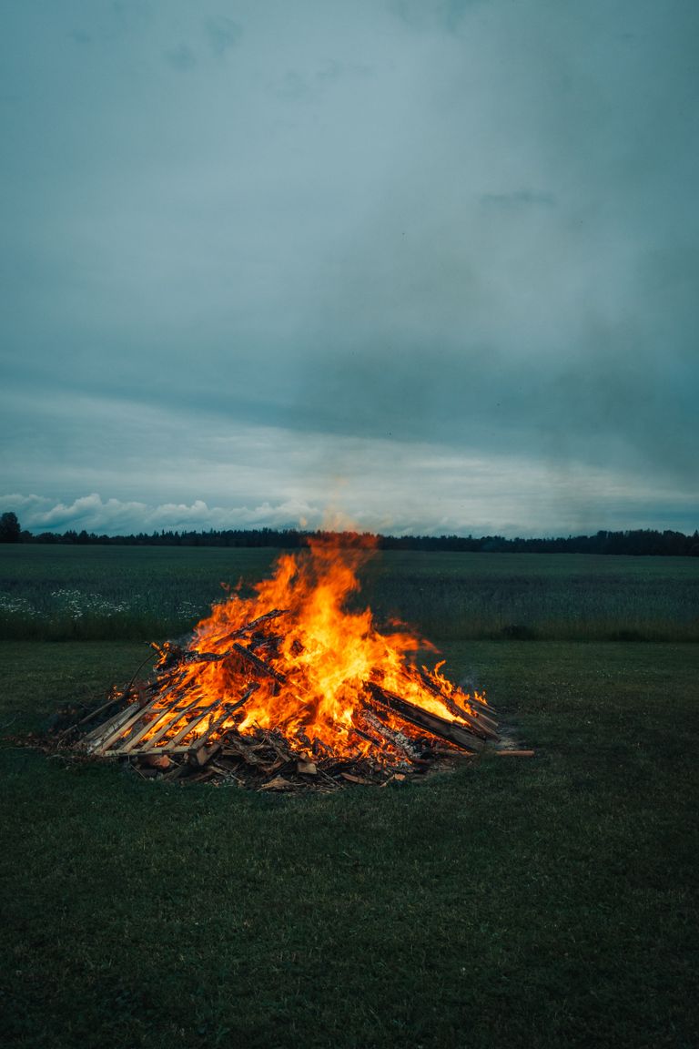 Large bonfire blazing in an open field, under a cloudy evening sky in rural setting.