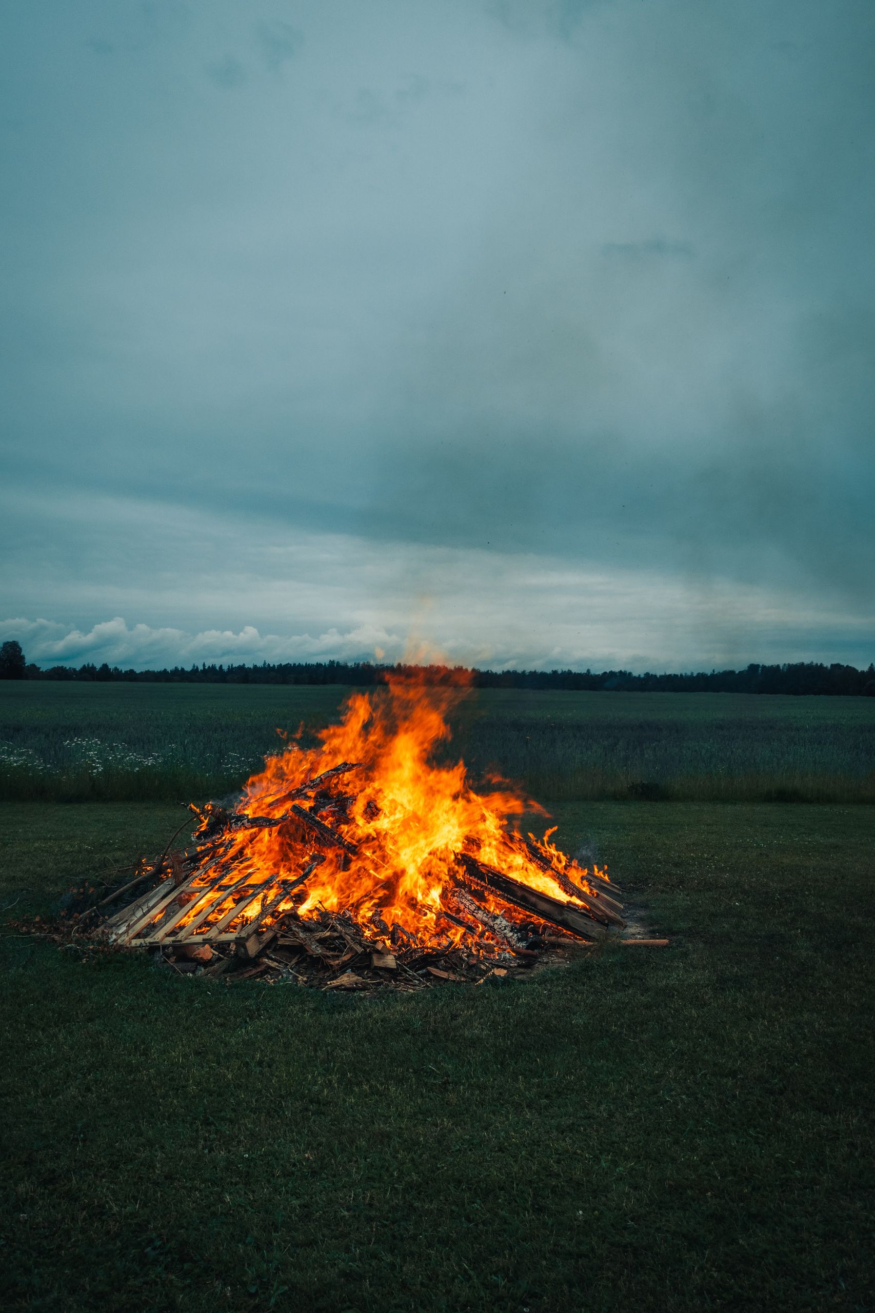 Large bonfire blazing in an open field, under a cloudy evening sky in rural setting.